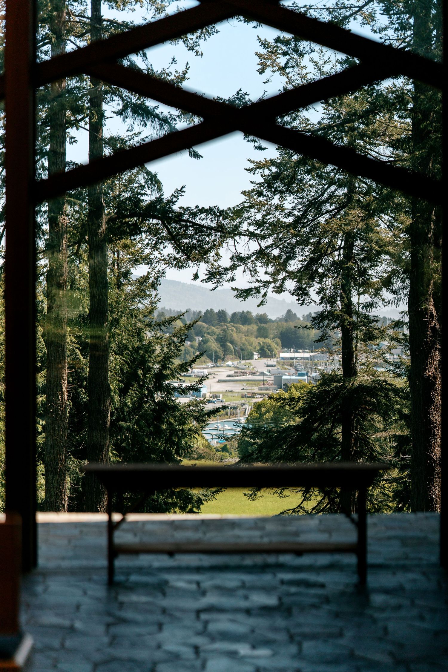 View through wooden chapel frame of tall pine trees and town below on clear day.