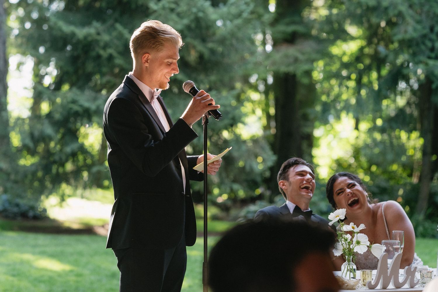 Groomsman delivering a heartfelt speech at microphone as bride and groom laugh at outdoor wedding.