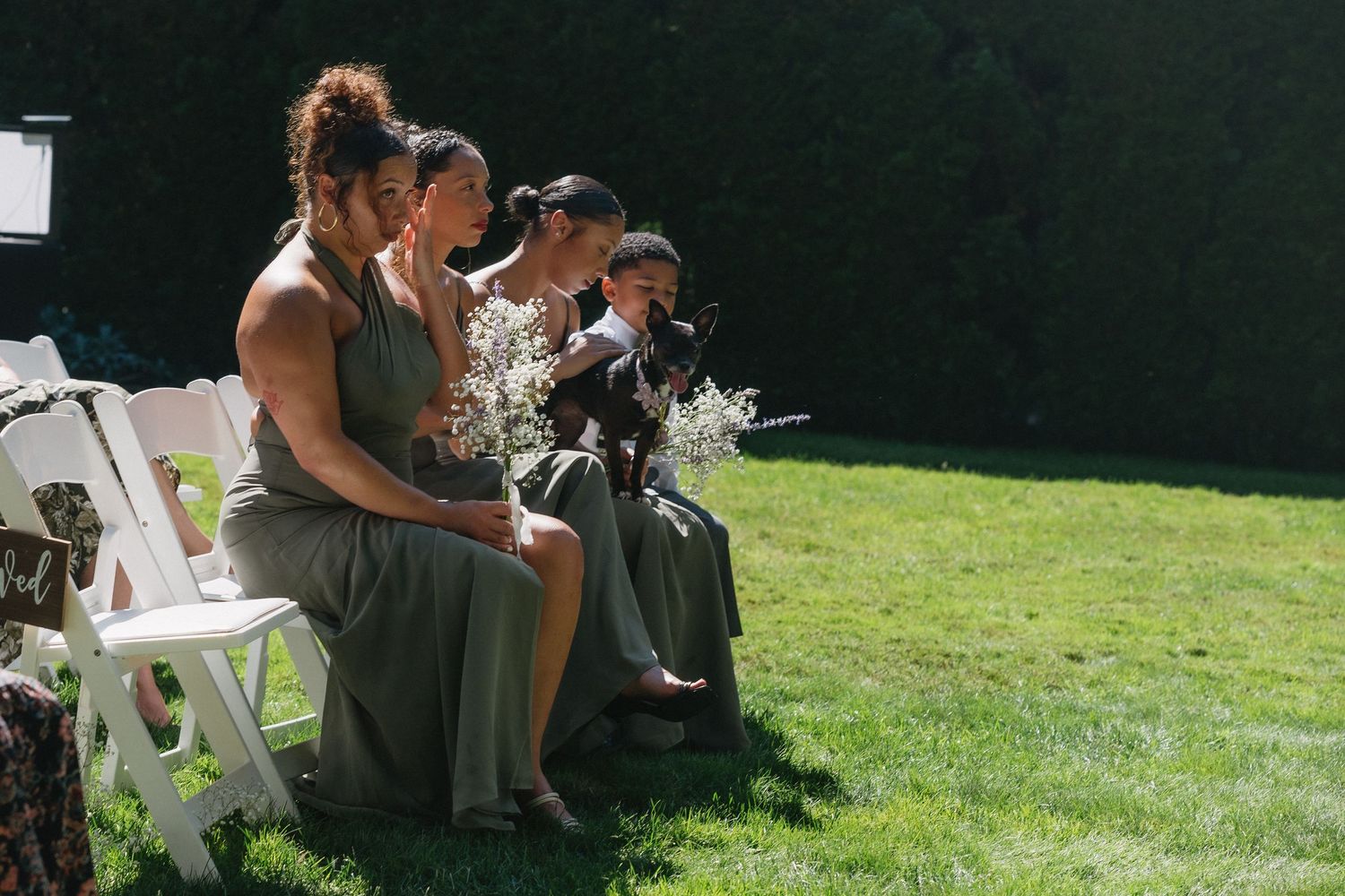 Bridesmaids in olive green dresses sit in white chairs holding floral bouquets at outdoor wedding ceremony.