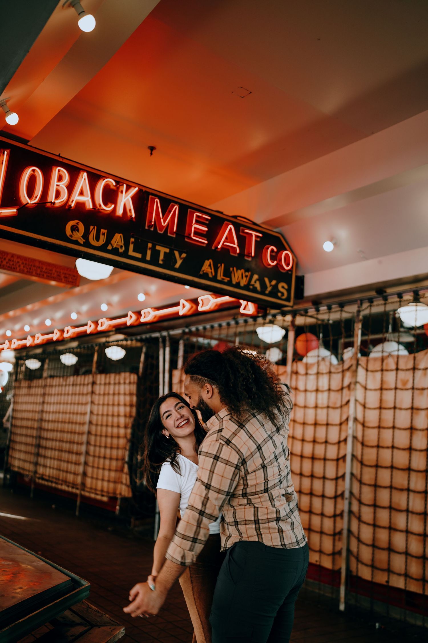 Laughing couple dancing inside Pike Place Market near Loback Meat Co neon sign.