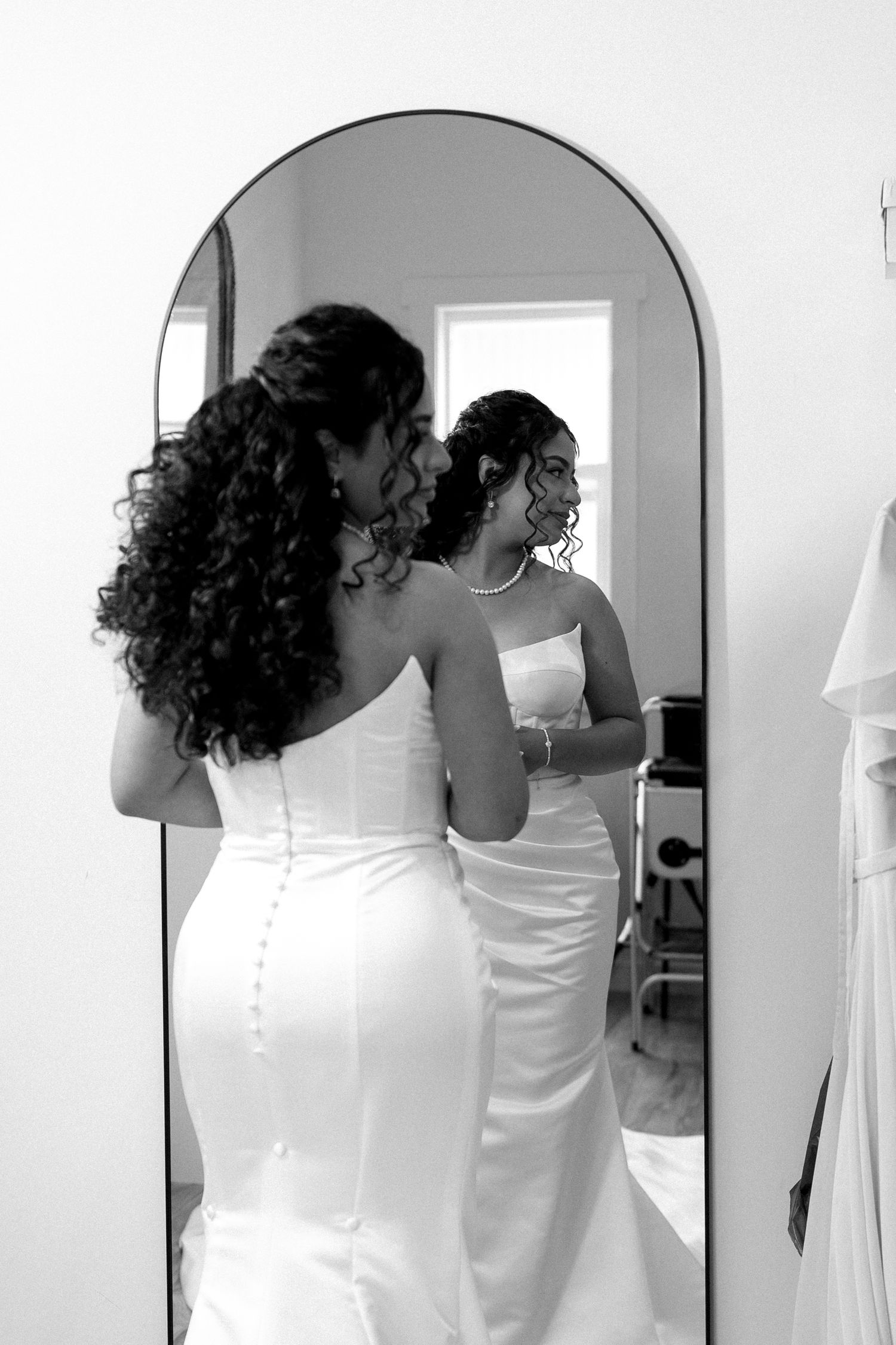 Black and white photo of bride in fitted strapless gown viewing reflection in arched mirror.