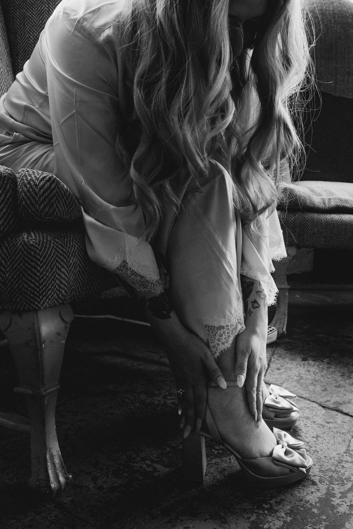 Black and white photo of bride putting on heeled wedding shoes while seated in vintage chair.