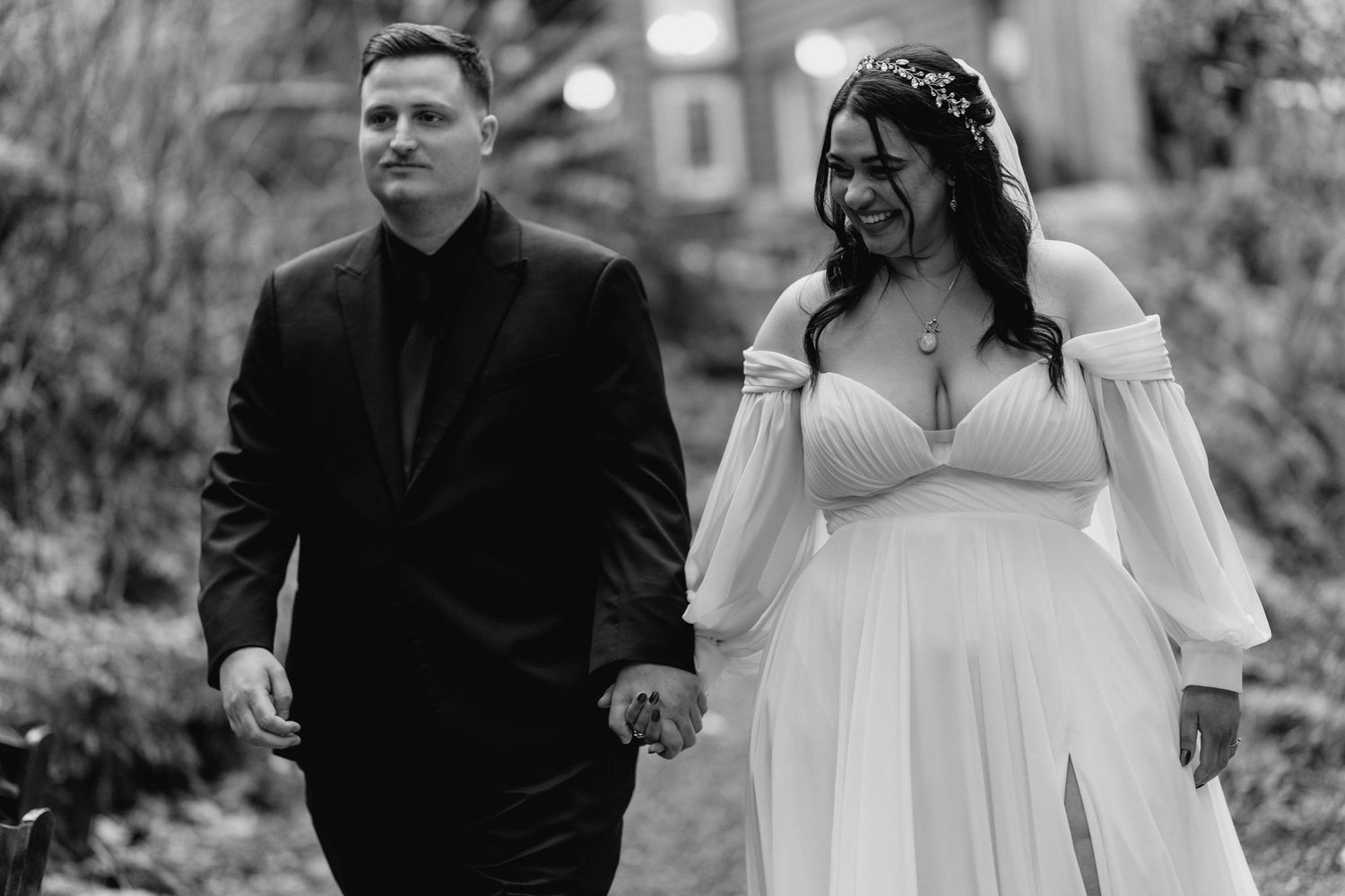Bride and groom hold hands walking together outdoors in black and white wedding portrait.
