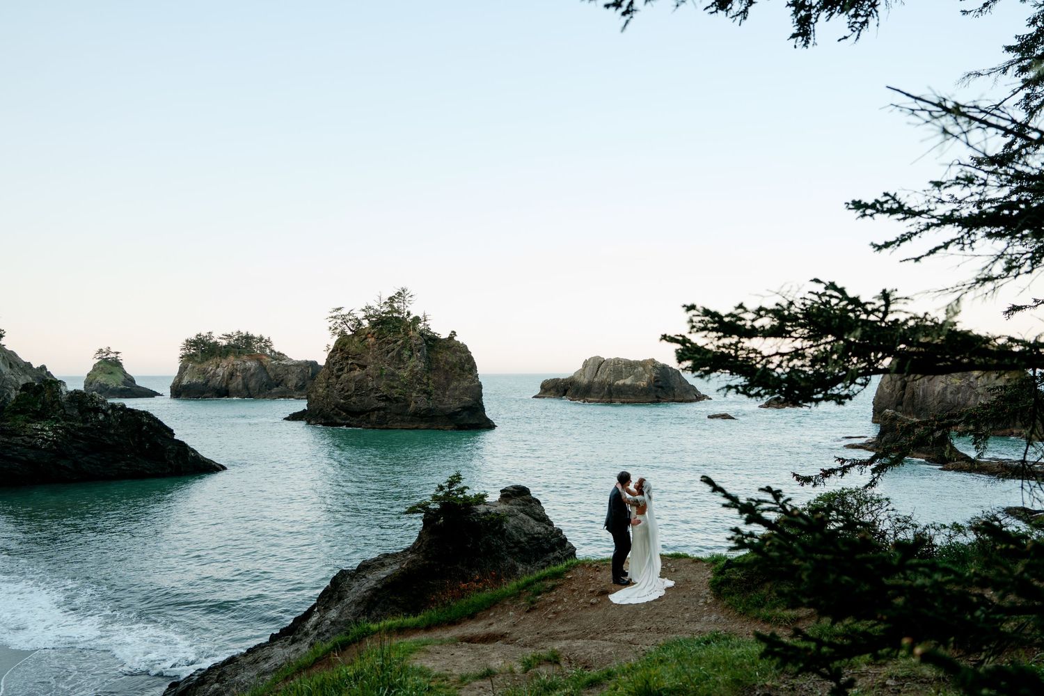 Wedding couple embraces on rugged Pacific coast cliff with scenic sea stacks rising from calm waters behind them.