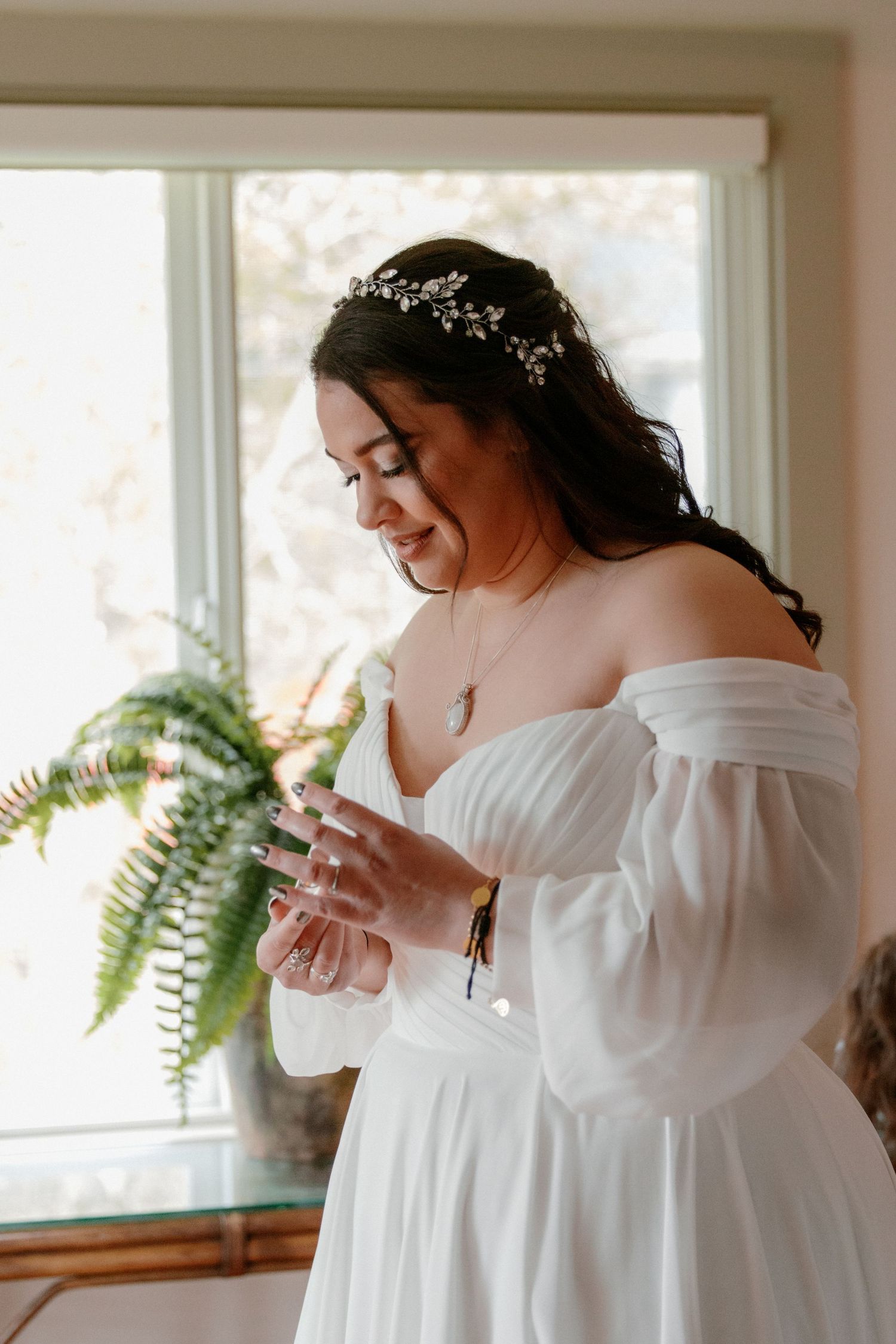 Smiling bride in off-shoulder white gown adjusting her sleeve, wearing a floral hair vine and pendant necklace.