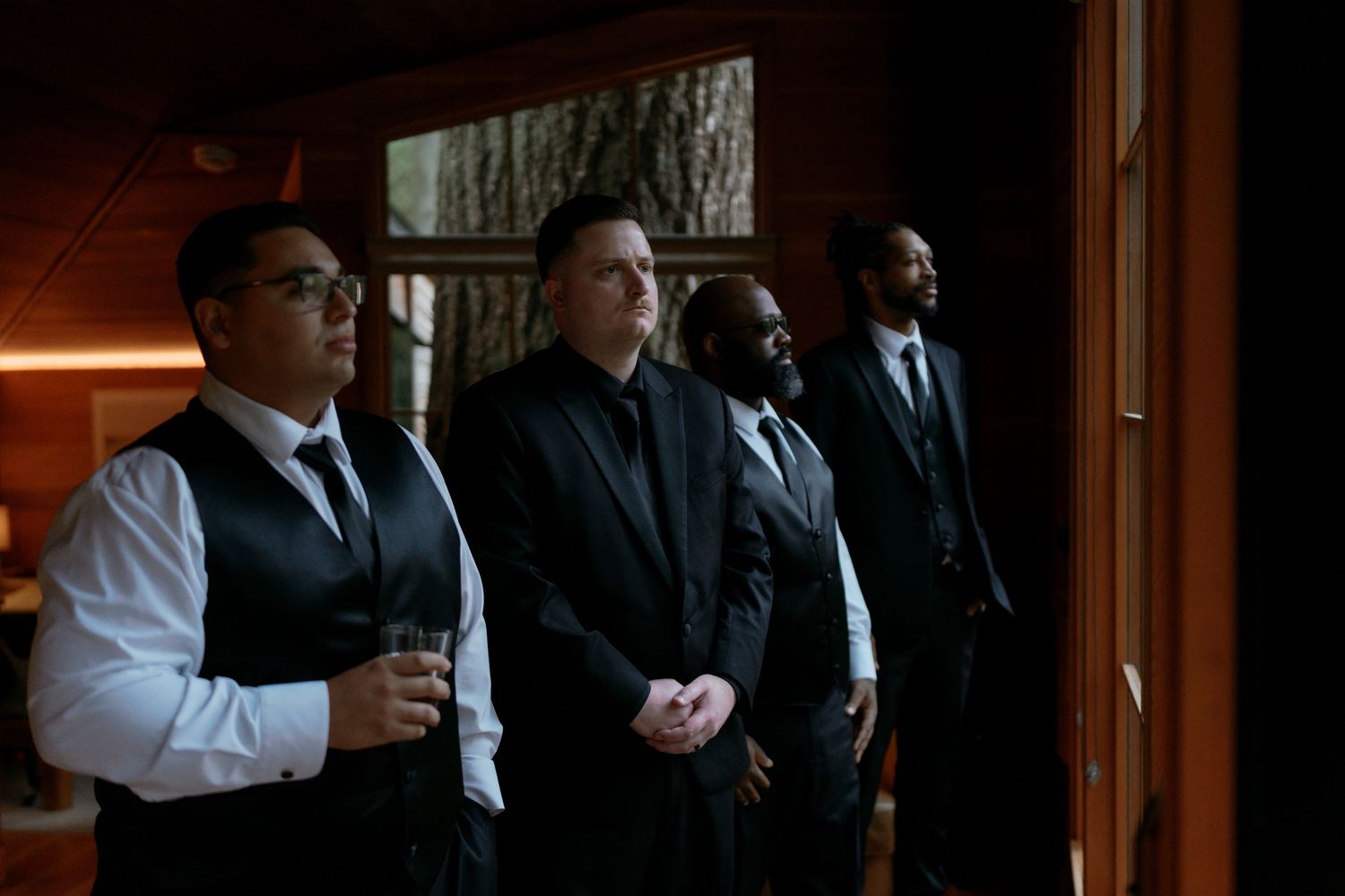 Four men in black suits and vests standing together indoors during wedding preparations.