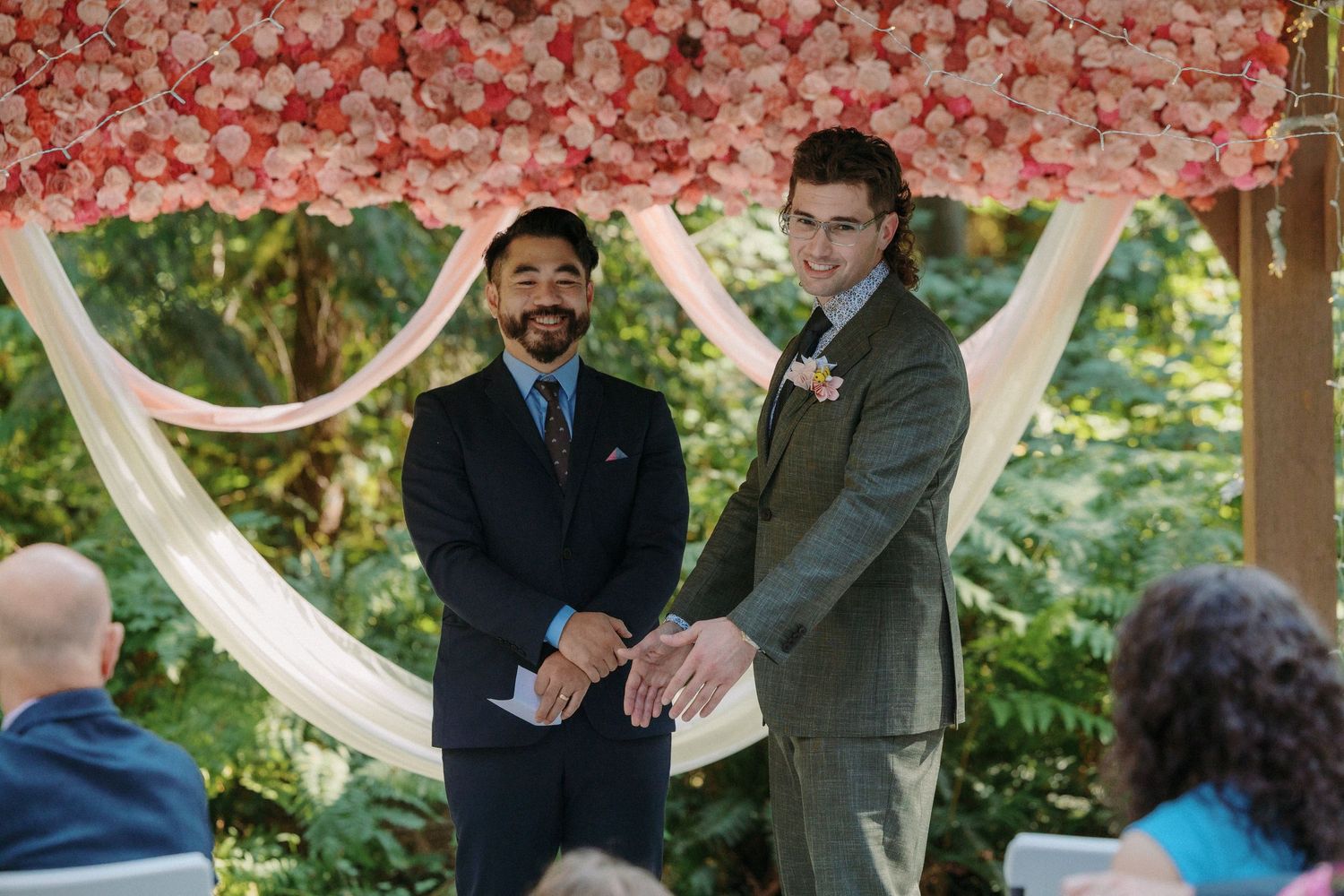 Two grooms smile holding hands at outdoor wedding altar adorned with pink floral arch amid lush greenery.