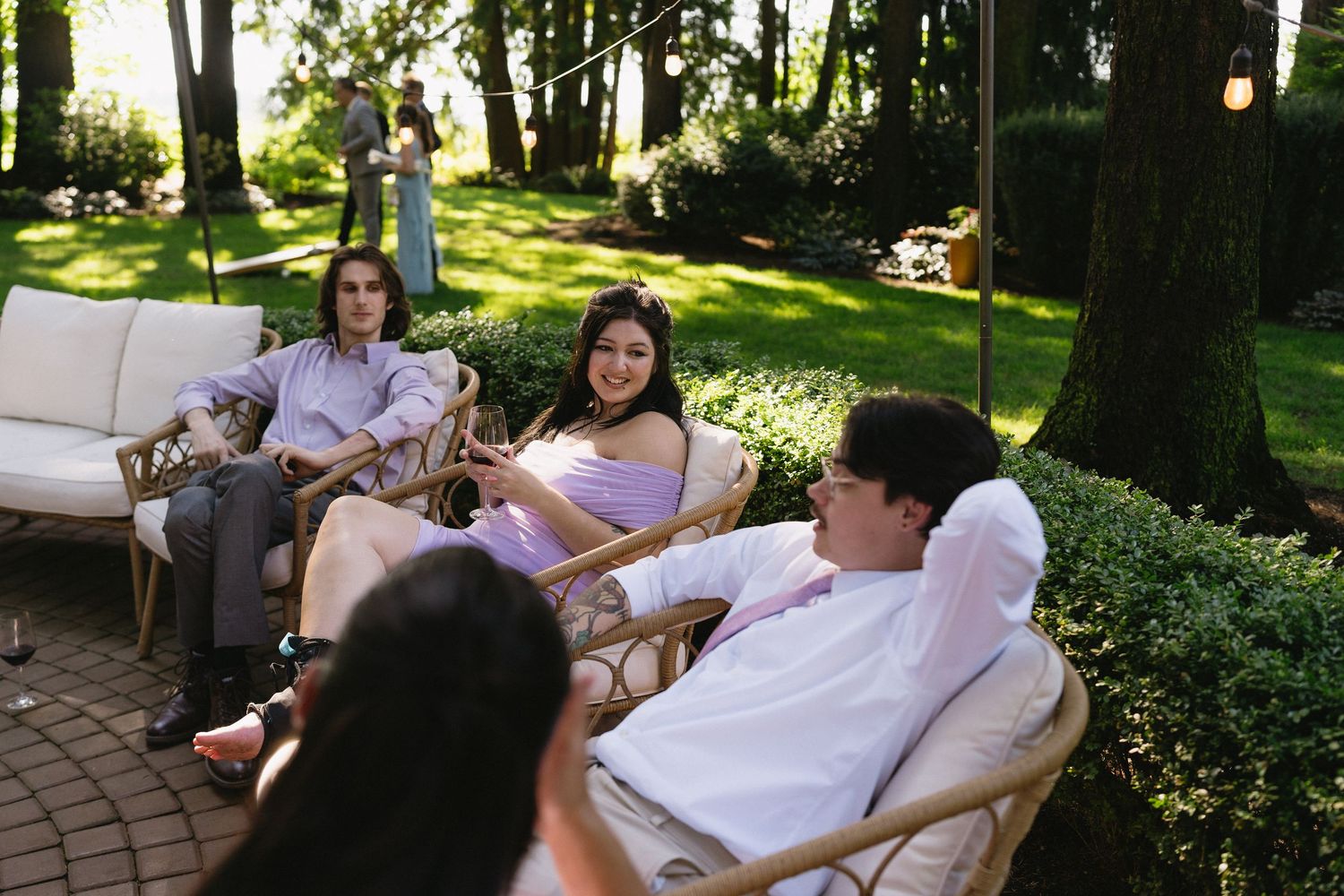 Wedding guests relax on outdoor patio furniture in lush green garden setting.