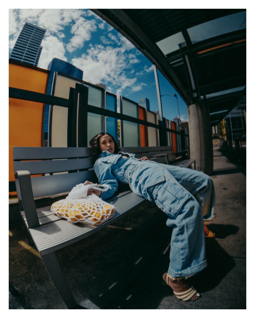 Woman in denim jumpsuit lounges on a bench at a colorful urban bus stop with a mesh bag nearby.
