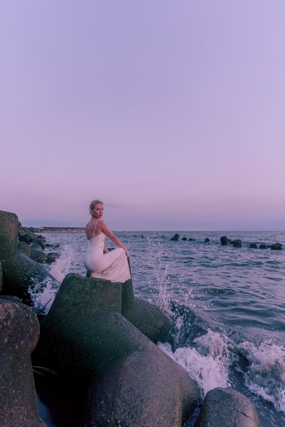 Blonde woman in white dress sits on coastal rocks with ocean waves at purple dusk.
