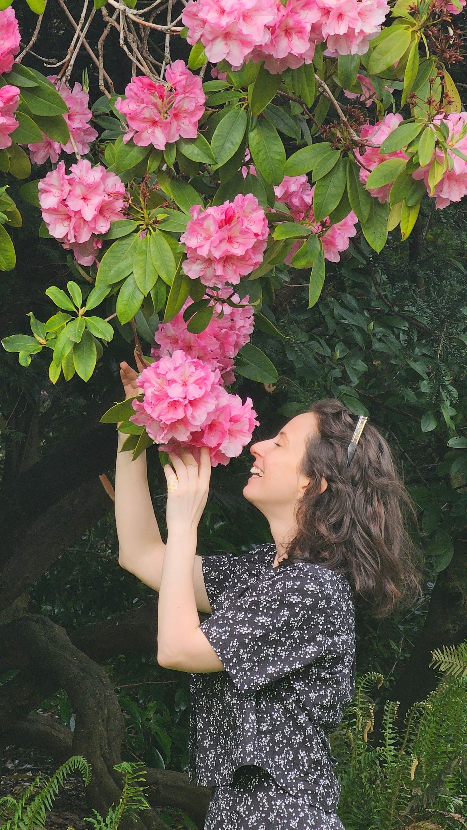 Smiling woman in floral dress joyfully smelling vibrant pink rhododendron blooms in a lush garden setting.