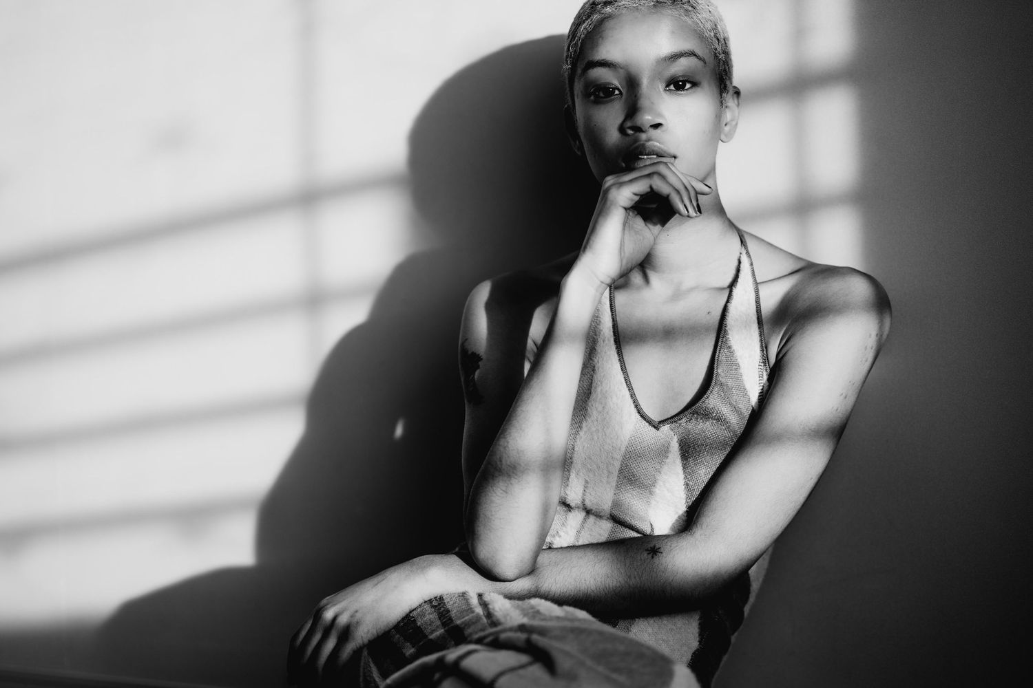 Black and white portrait of a woman sitting thoughtfully with dramatic shadow patterns on the wall behind her.