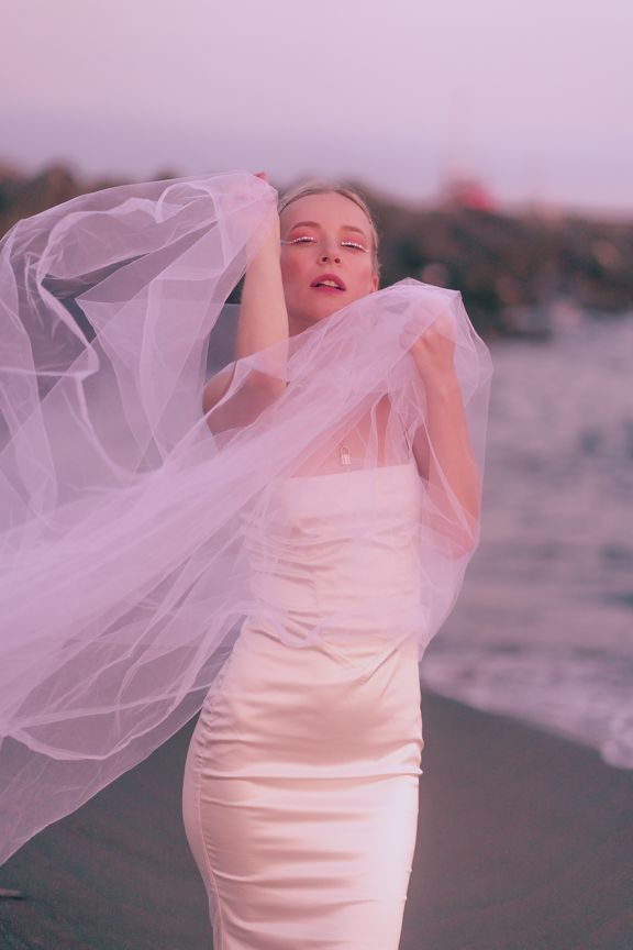 Blonde bride in white dress holds billowing tulle veil on rocky beach at dusk.