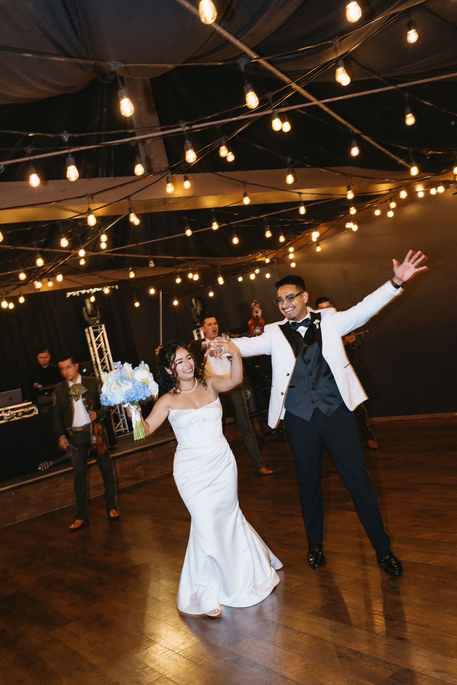 Bride and groom sharing joyful first dance under warm string lights at reception.