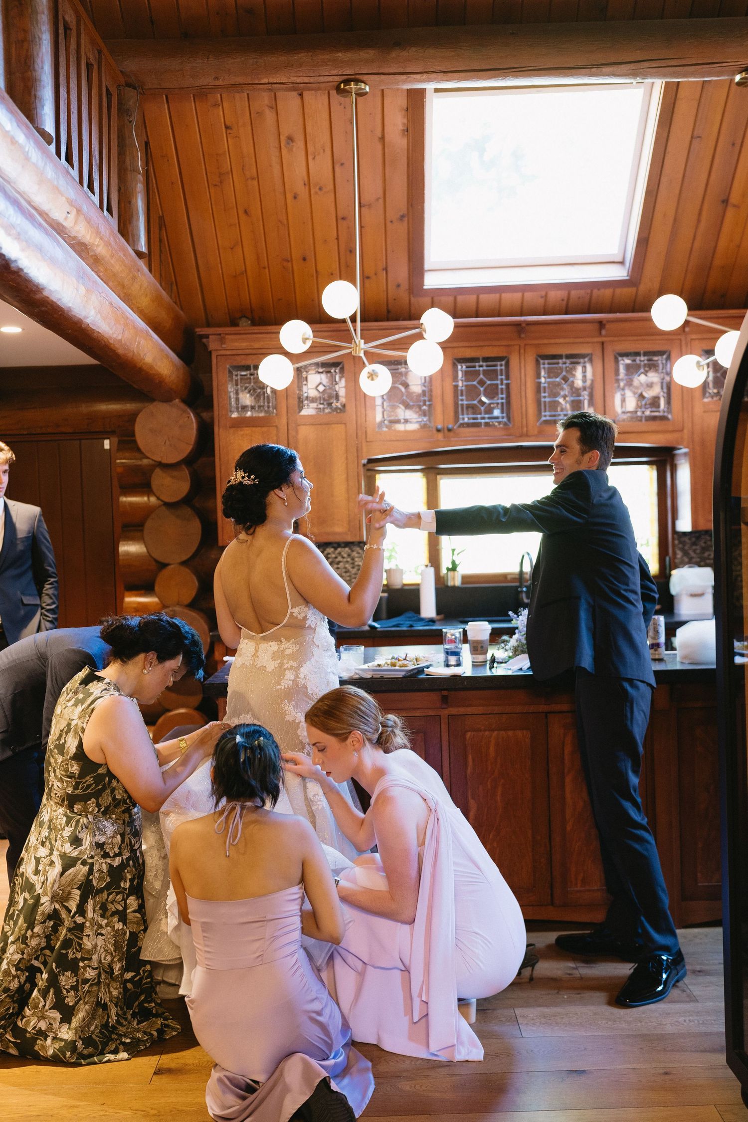 Bridesmaids help bride with dress while groom stands at bar in rustic wood cabin wedding venue.