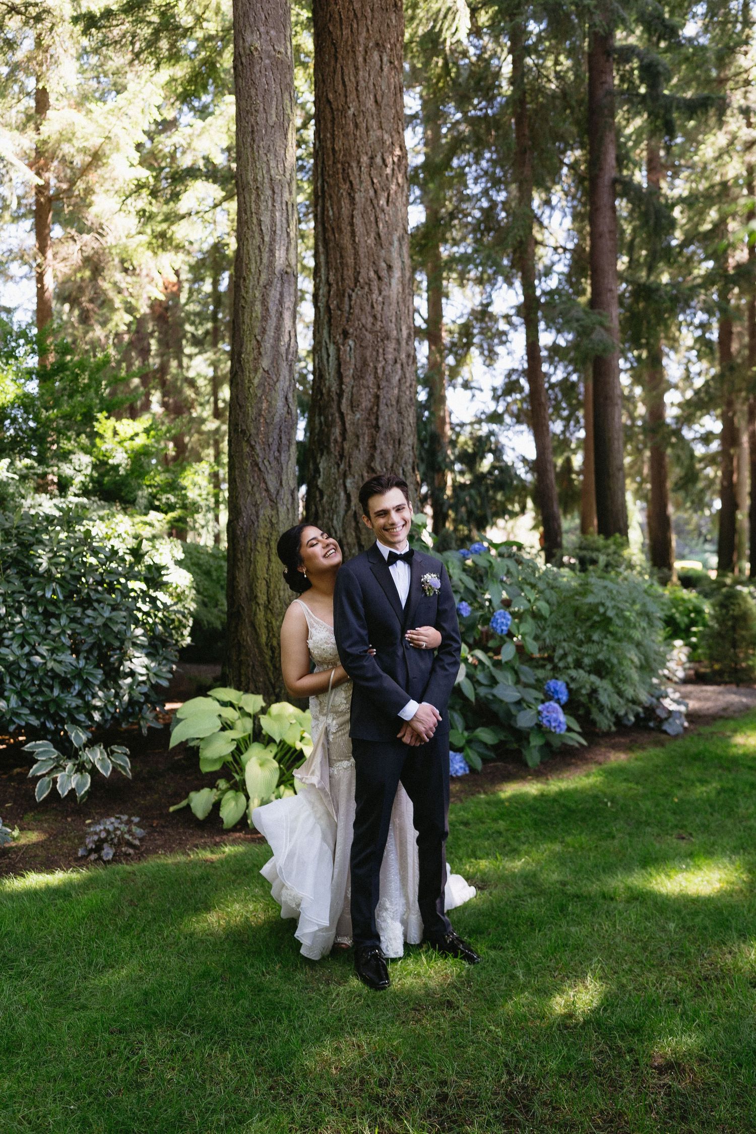 Bride in lace mermaid gown and groom in black tuxedo posing among tall pine trees outdoors.