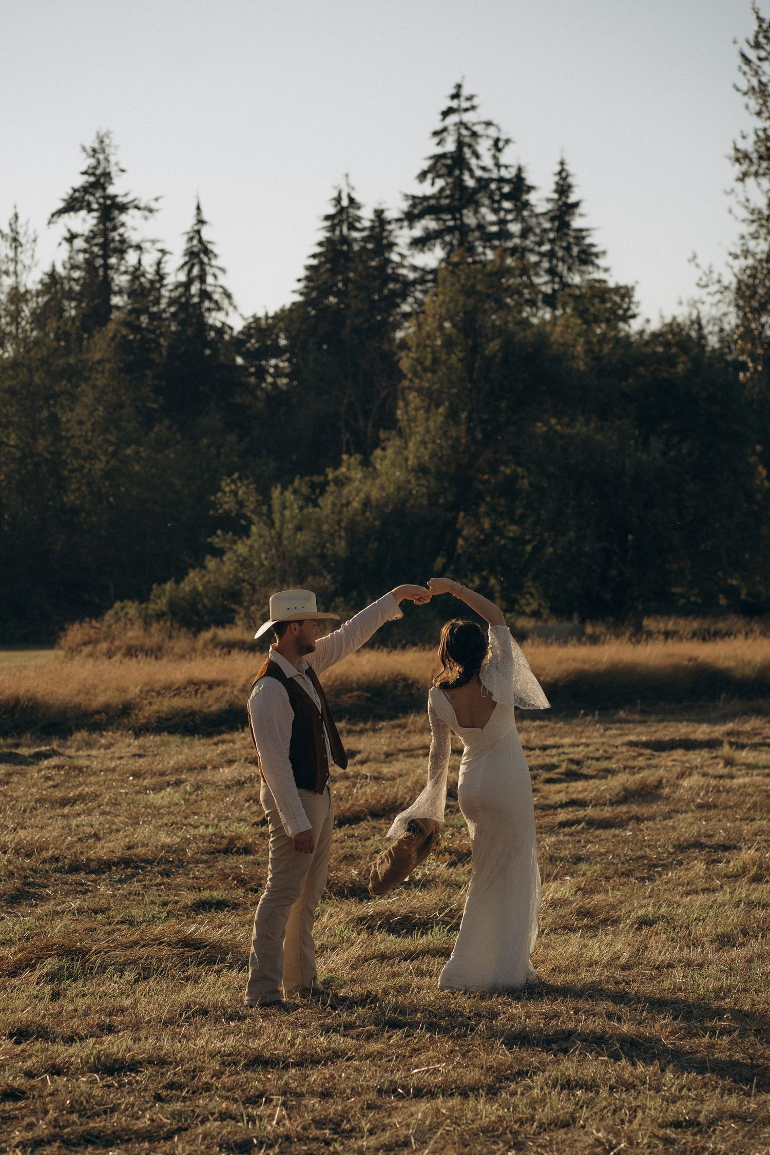 Bride and groom dancing together in rustic field at golden hour with evergreen backdrop.