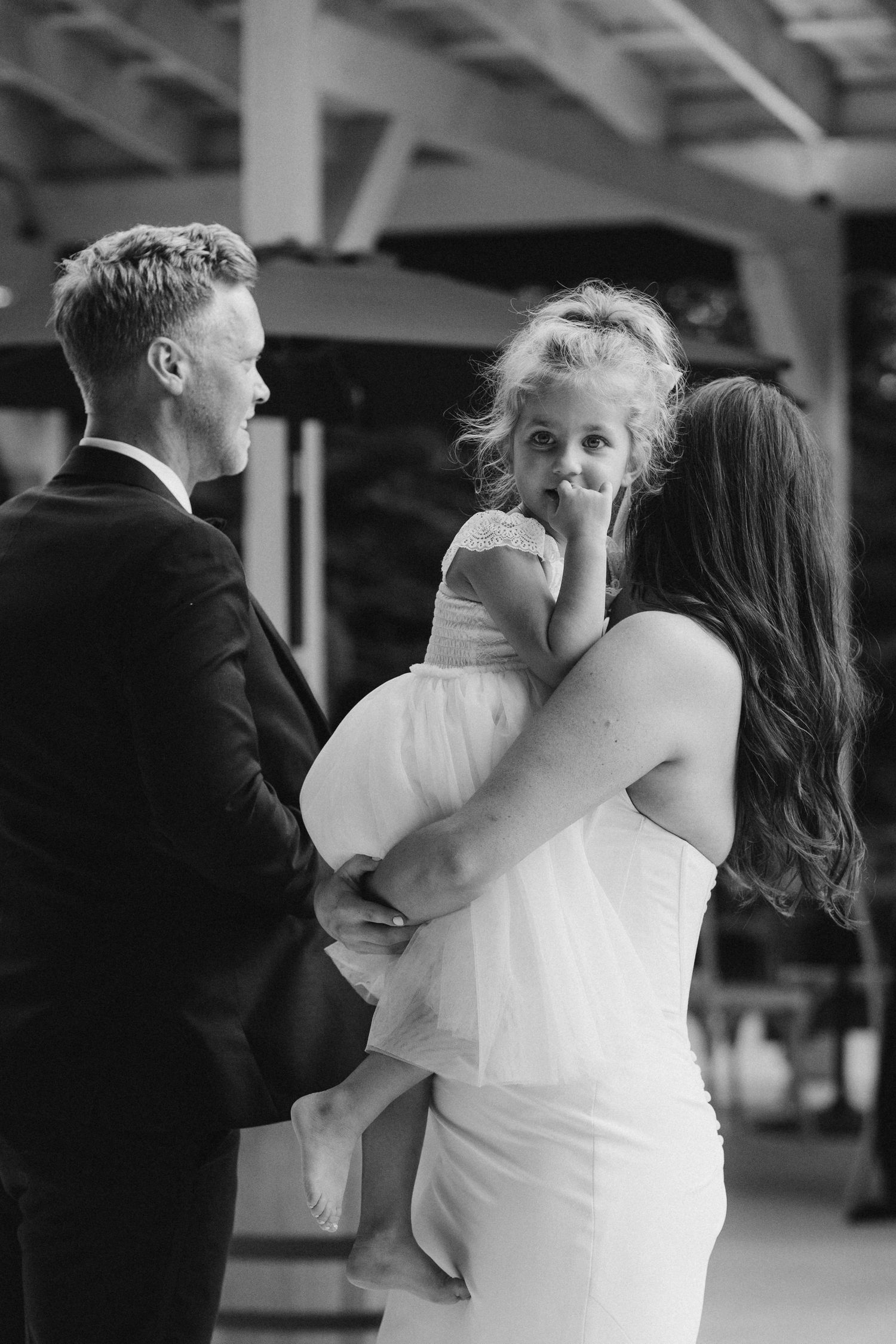Black and white photo of bride and groom holding a young flower girl at wedding reception.