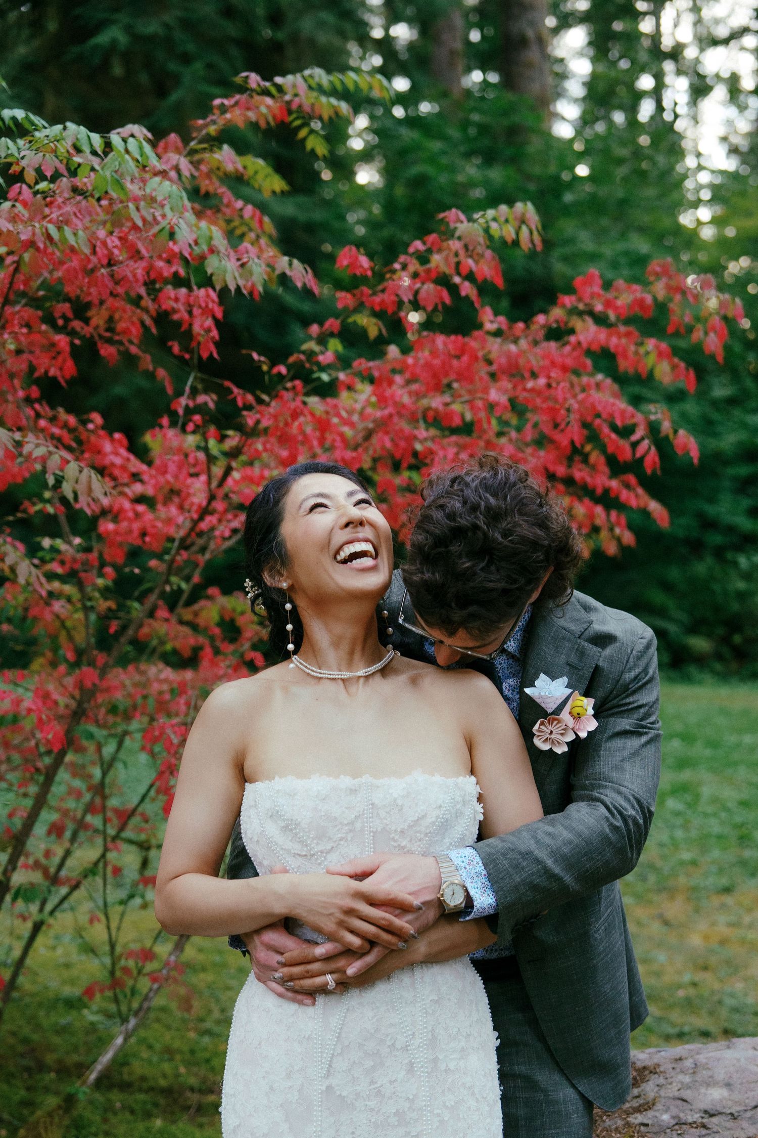 Bride laughs joyfully as groom embraces her near vibrant red autumn foliage in an outdoor wedding photo.