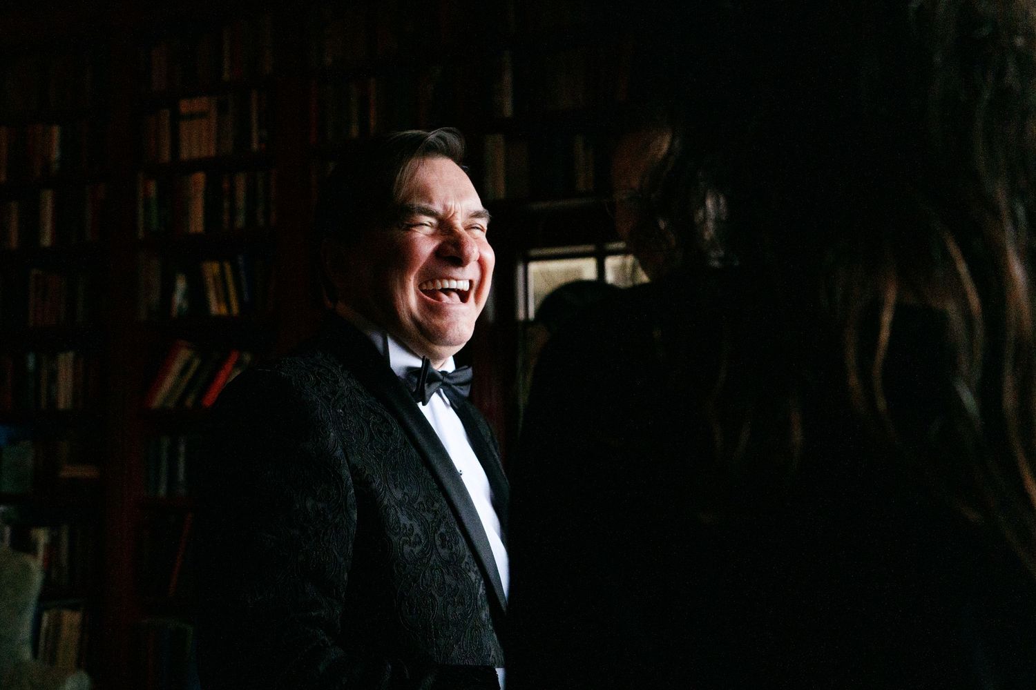 Man in black tuxedo laughing joyfully in dimly lit library setting.