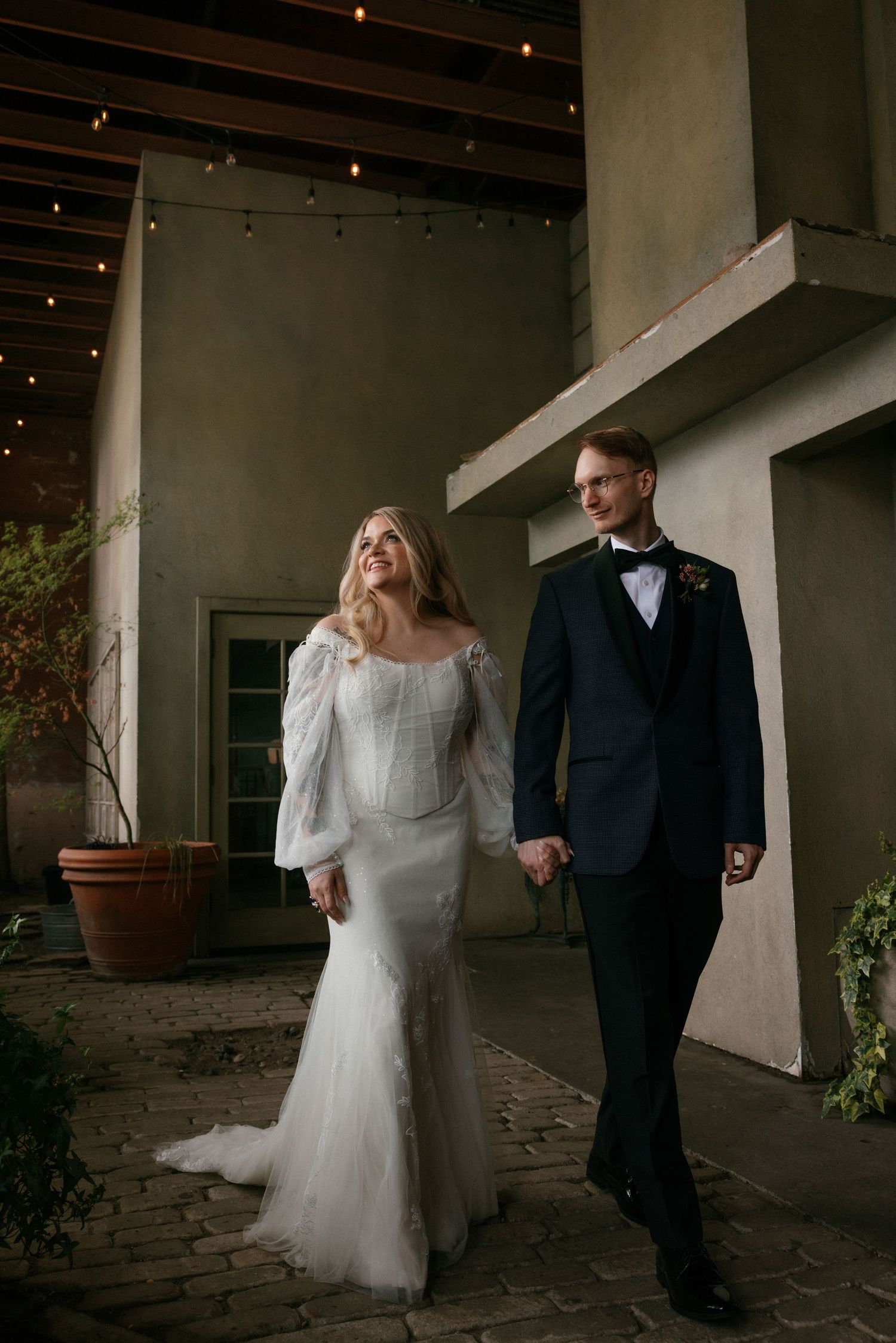 Bride and groom walking hand in hand under string lights on a covered outdoor patio at night.