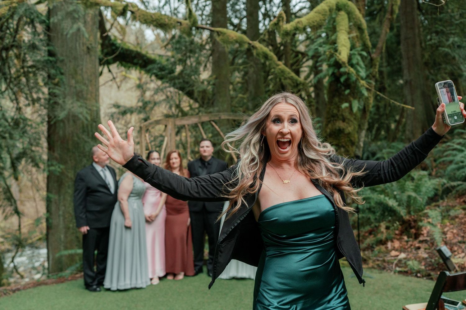 Excited blonde woman in teal dress celebrating with arms raised at outdoor wedding reception in forest.