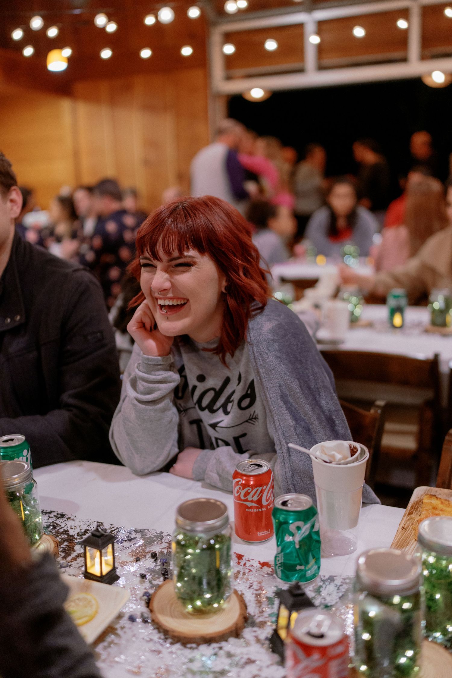 Laughing red-haired wedding guest in casual attire enjoys the reception at a table with mason jar centerpieces.