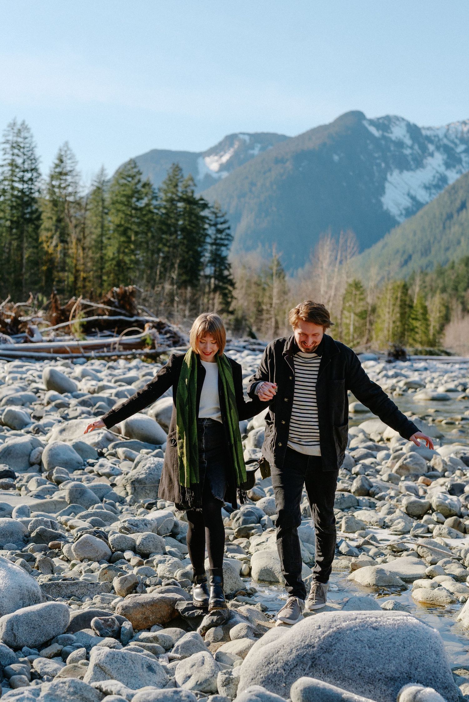 Couple balancing on rocky riverbed with snow-capped mountains and evergreen trees in background.