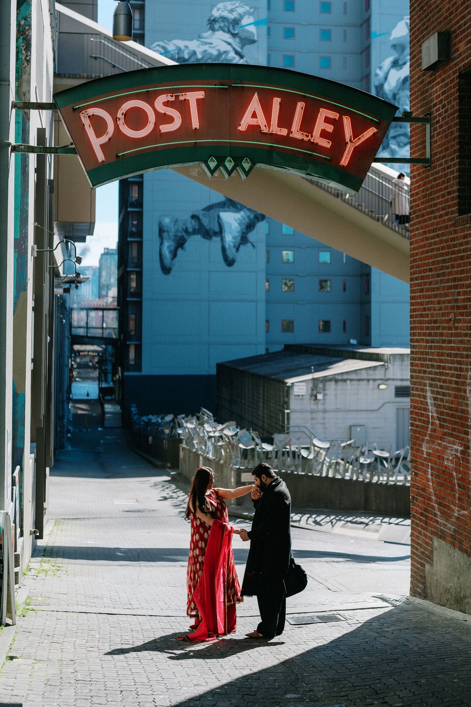 Couple in traditional Indian attire standing under Seattle's Post Alley neon sign in warm sunlight.