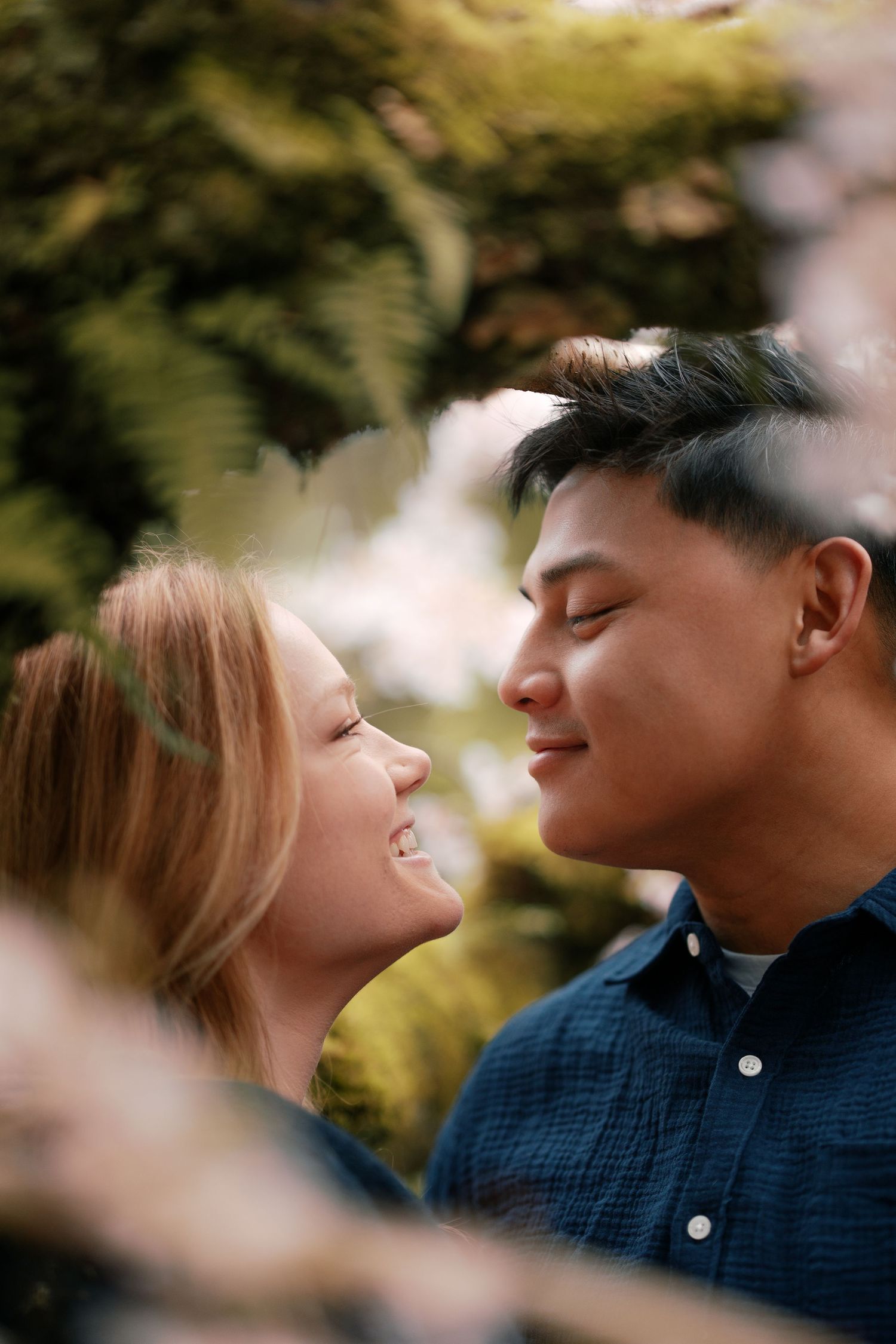 Close-up of smiling couple nearly kissing surrounded by soft pink cherry blossoms.