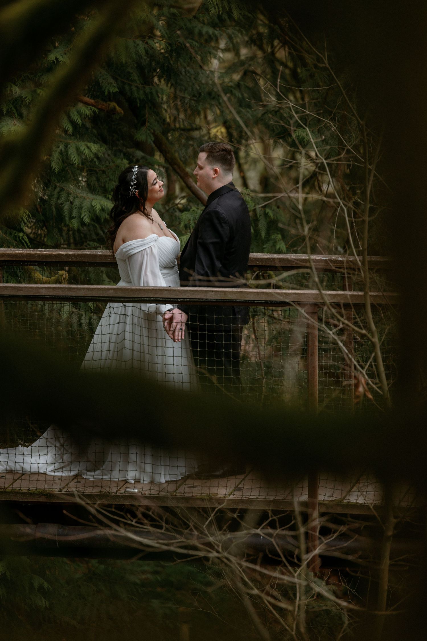 Bride in white off-shoulder gown and groom in black suit gaze at each other on forest bridge.