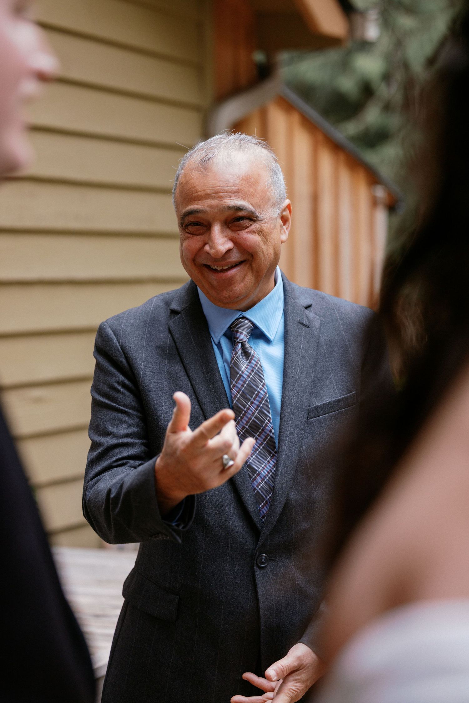 Older gentleman in gray suit smiling warmly while gesturing at an outdoor wedding event.