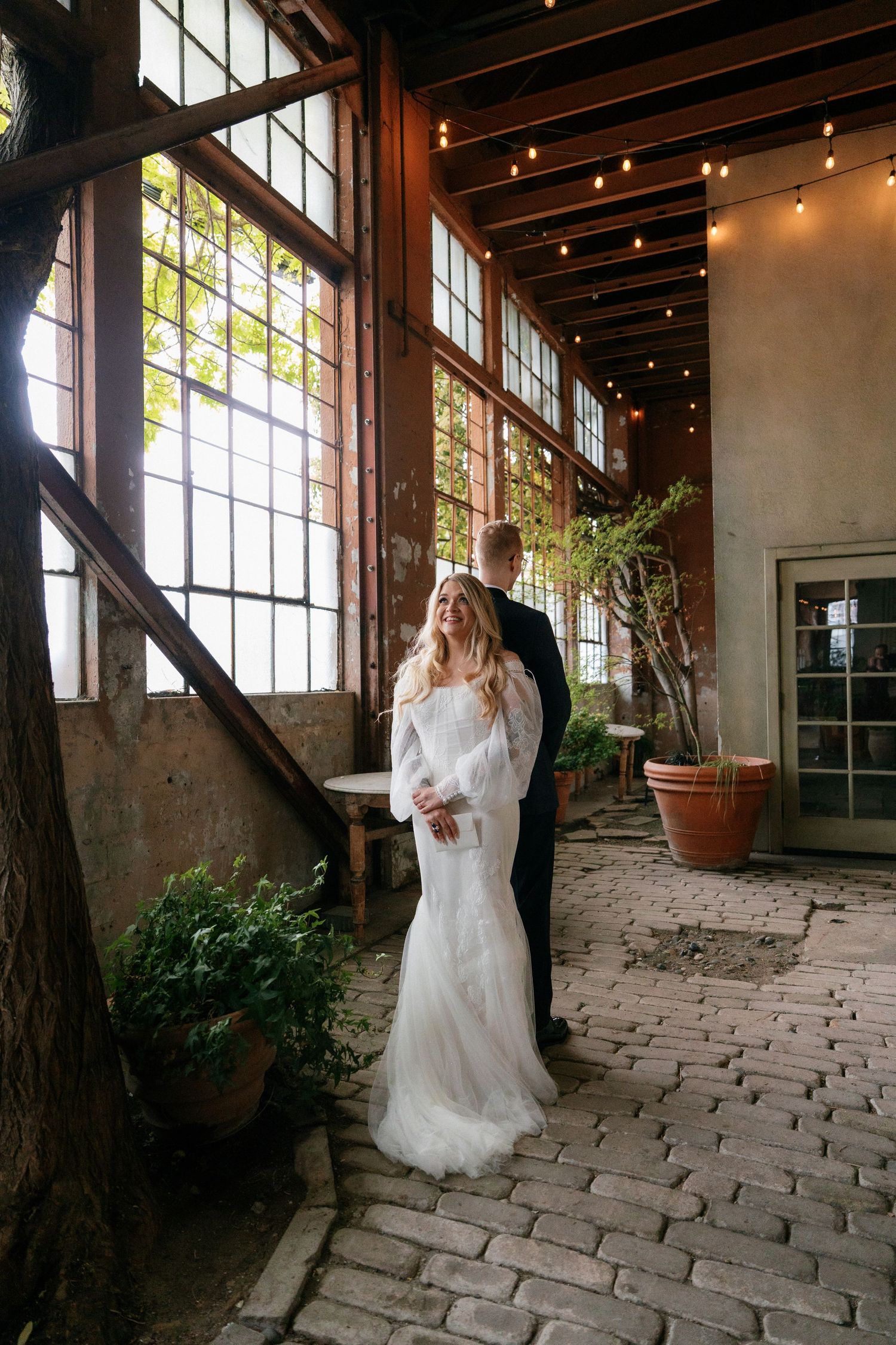 Bride and groom standing together in a rustic industrial venue with cobblestone floors and string lights.