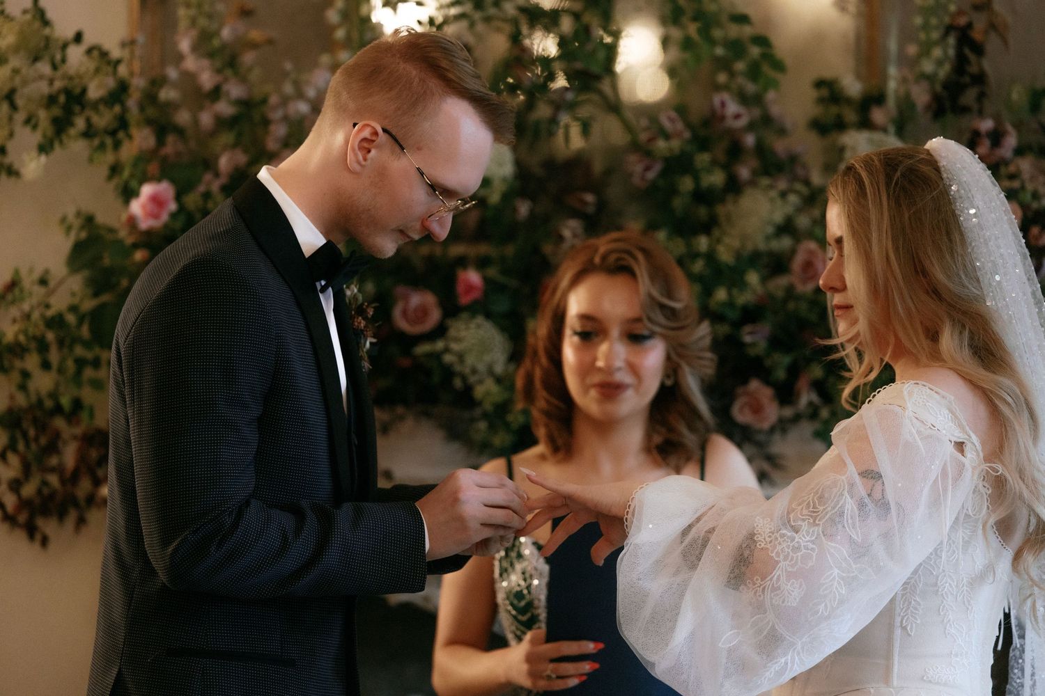 Groom places wedding ring on bride's finger assisted by bridesmaid at indoor ceremony.