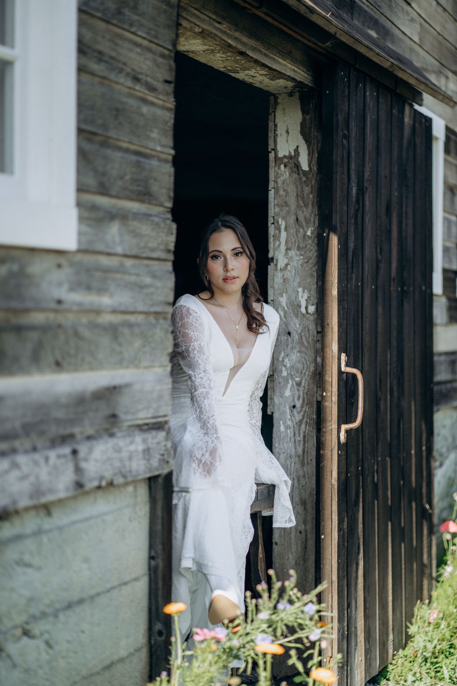 Elegant bride in white lace bell-sleeve gown standing in weathered wooden barn doorway with wildflowers.