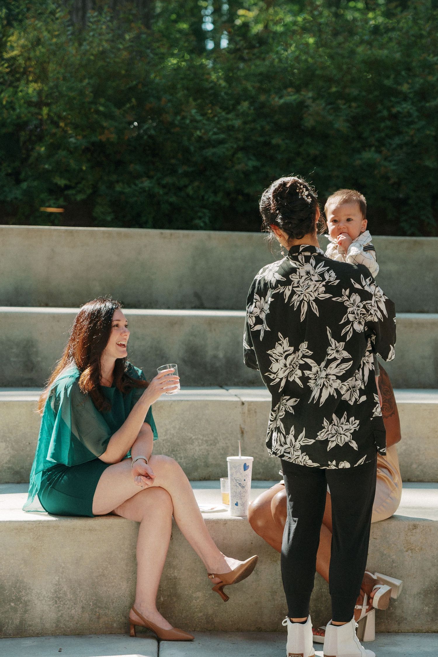 A woman in a green dress holds a drink while chatting with a person carrying a baby outdoors.