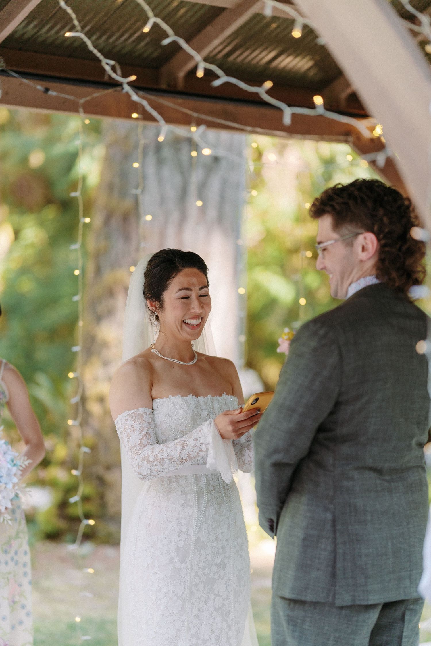 Laughing Asian bride in off-shoulder lace gown reads vows to groom under string light canopy.