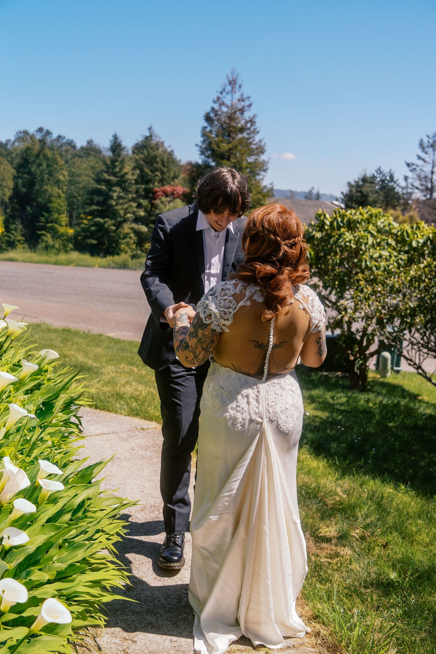 Groom in black suit playfully dances with tattooed bride in lace wedding dress along a sunny garden path.