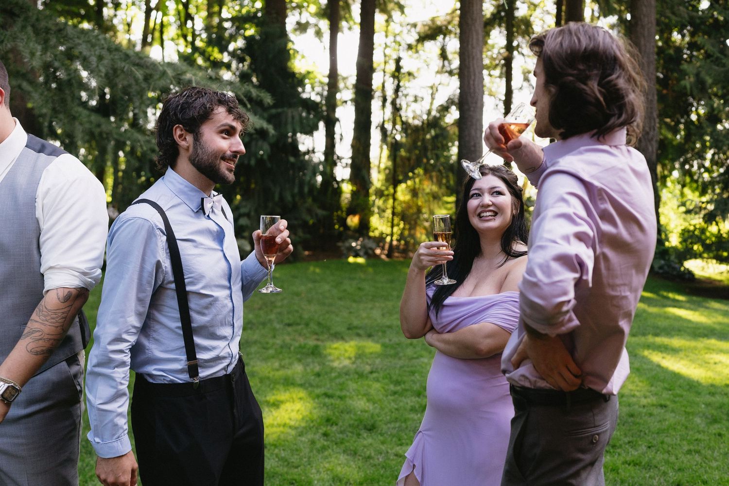 Wedding guests laughing and enjoying drinks outdoors in a sunlit garden with tall trees.