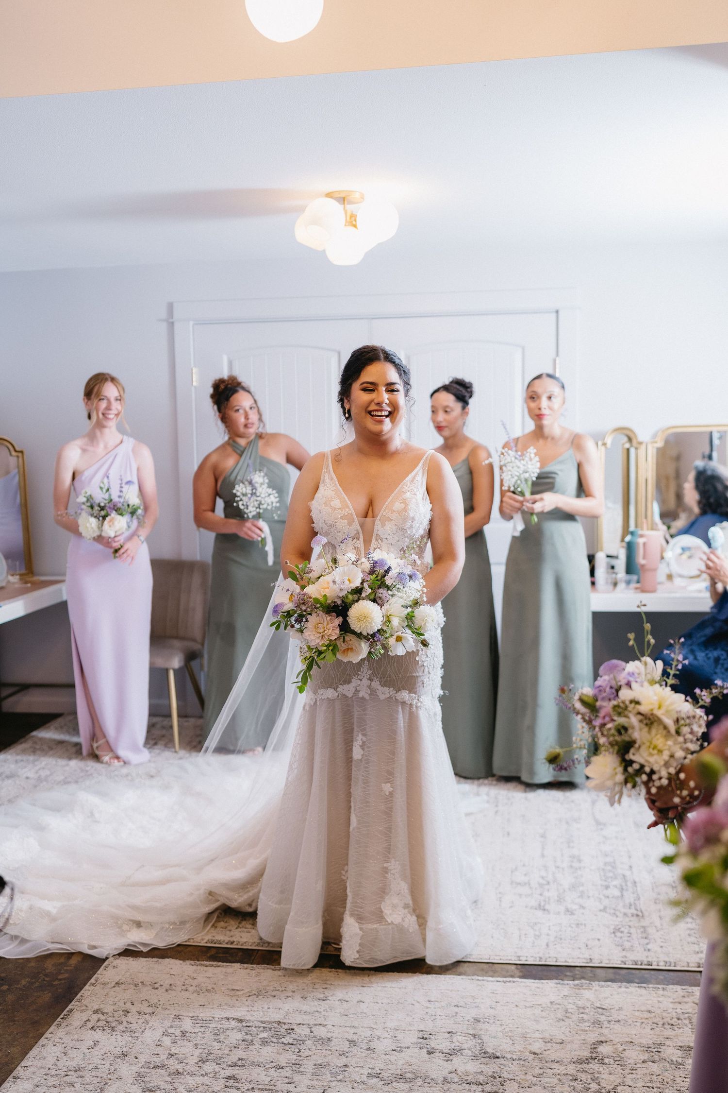 Smiling bride holding bouquet in lace gown with bridesmaids in sage and lavender behind her.