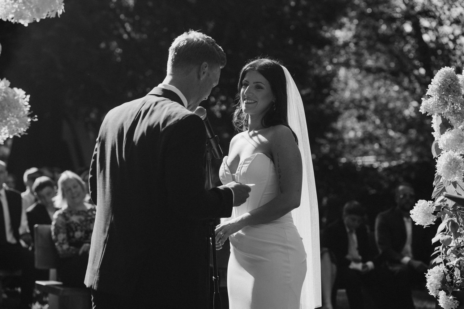 Black and white photo of bride and groom exchanging vows at outdoor wedding ceremony.