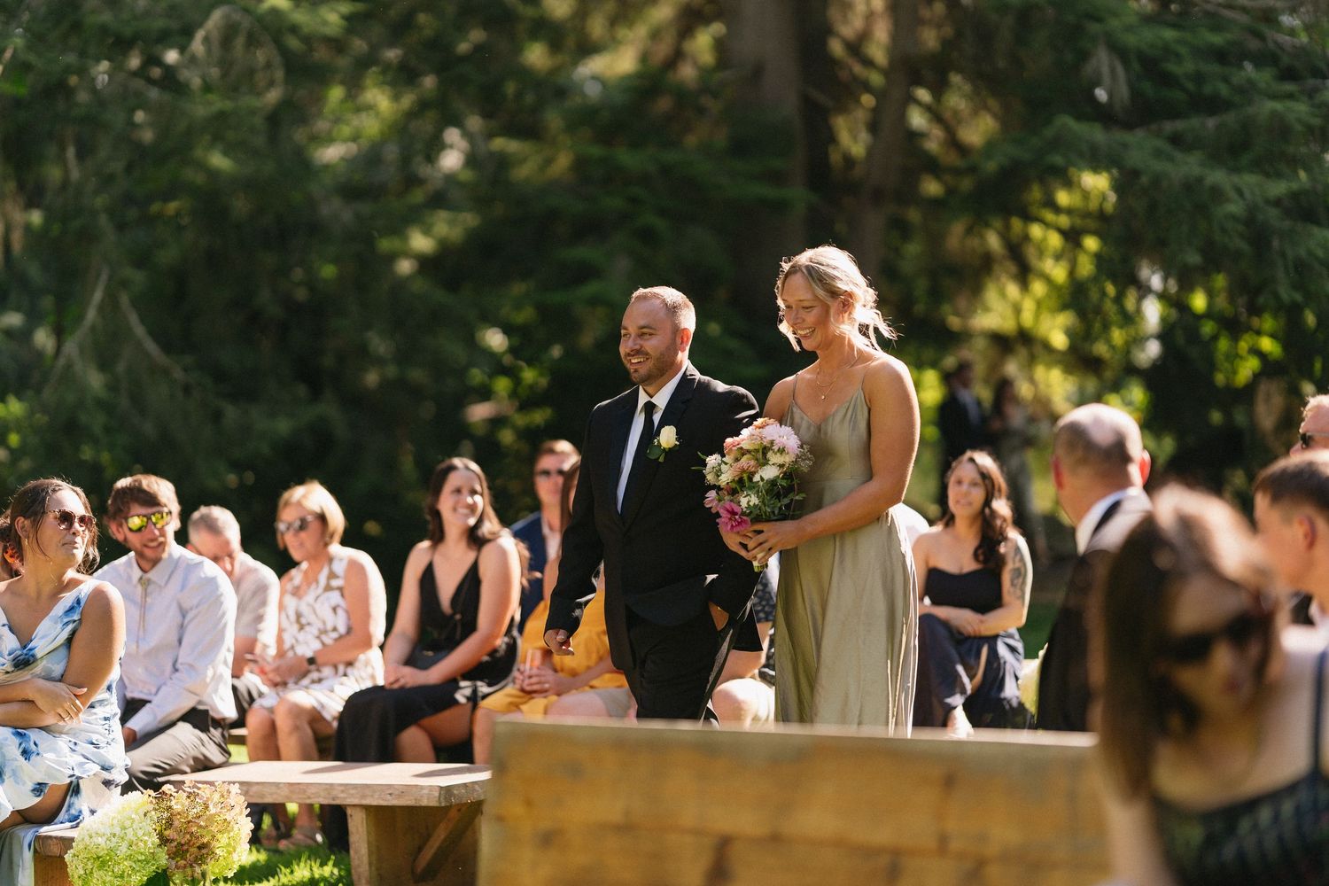 Bridesmaid in champagne dress walks down outdoor wedding aisle holding floral bouquet with guests seated.