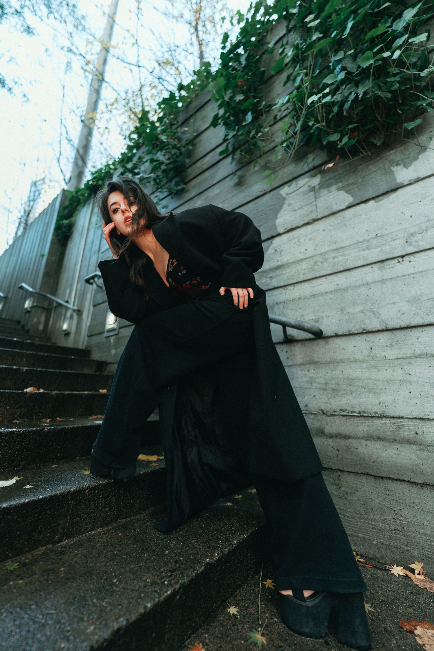 Woman in dramatic black coat and wide-leg pants poses on outdoor stairs with ivy-covered wall.