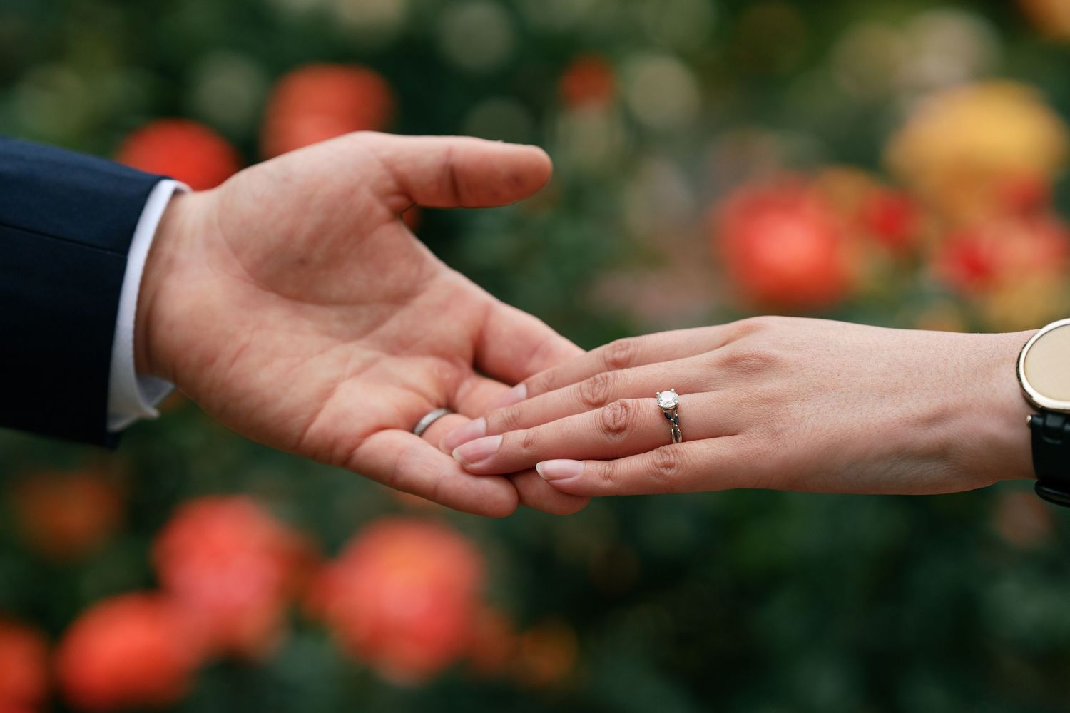 Close-up of bride and groom holding hands displaying wedding rings with orange roses background.