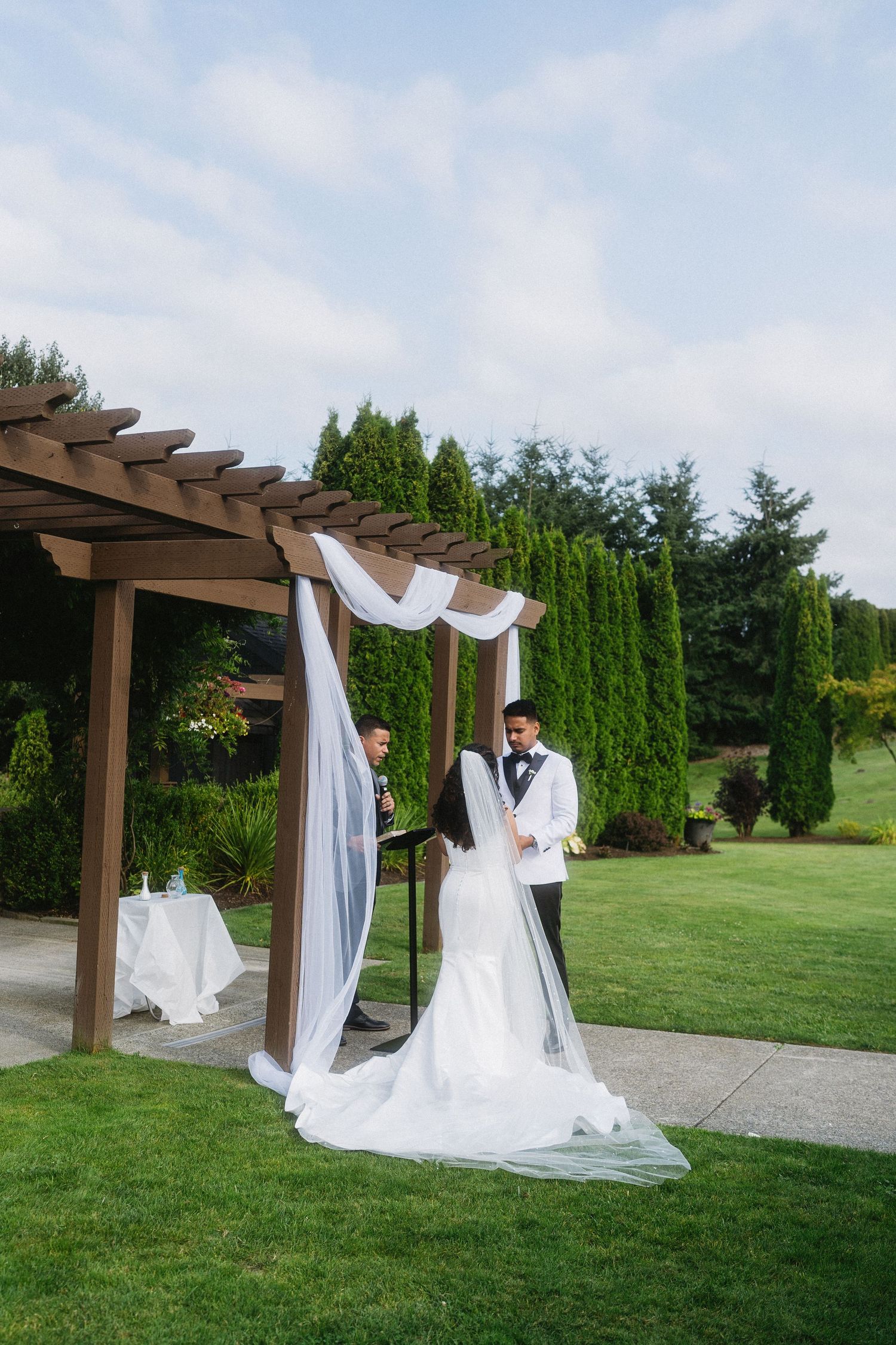 Bride and groom stand under a draped wooden pergola during an outdoor wedding ceremony on a lush green lawn.
