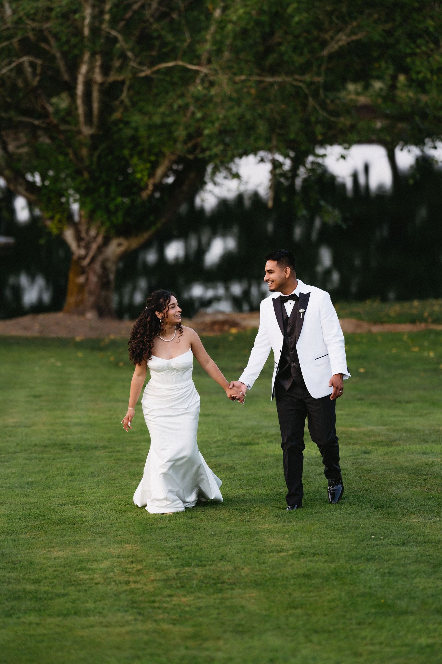 Bride and groom walk hand-in-hand across a green lawn, smiling at each other on their wedding day.