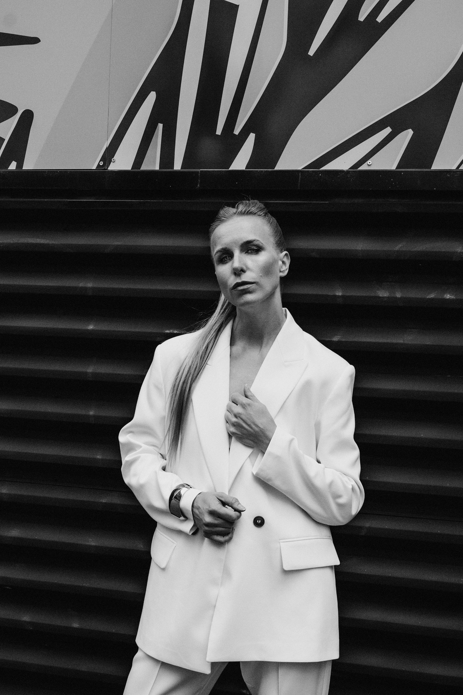 Blonde woman in white double-breasted blazer poses against dark corrugated wall in black and white fashion portrait.