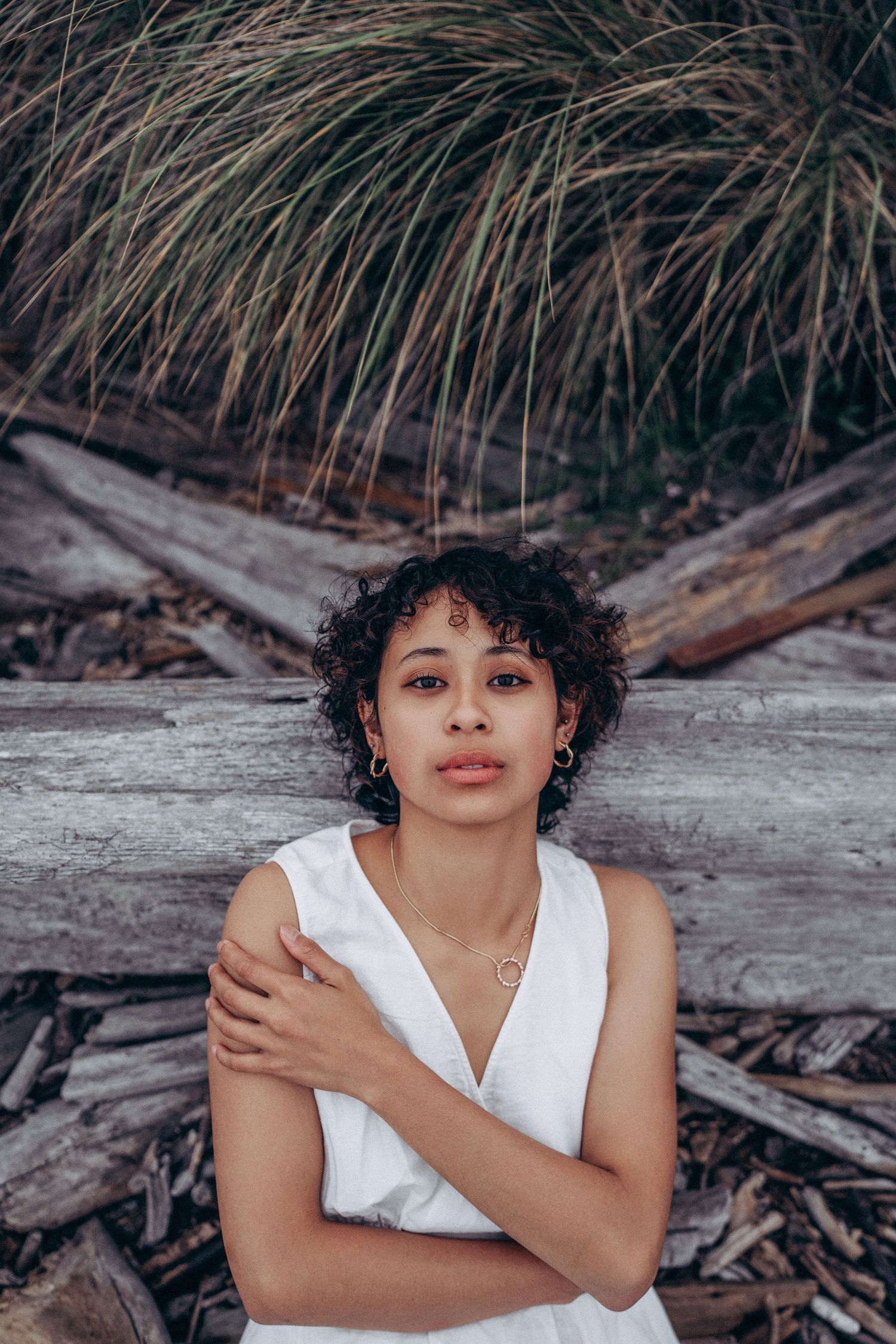 Woman with curly hair in white dress crosses arms standing before driftwood and tall grass.