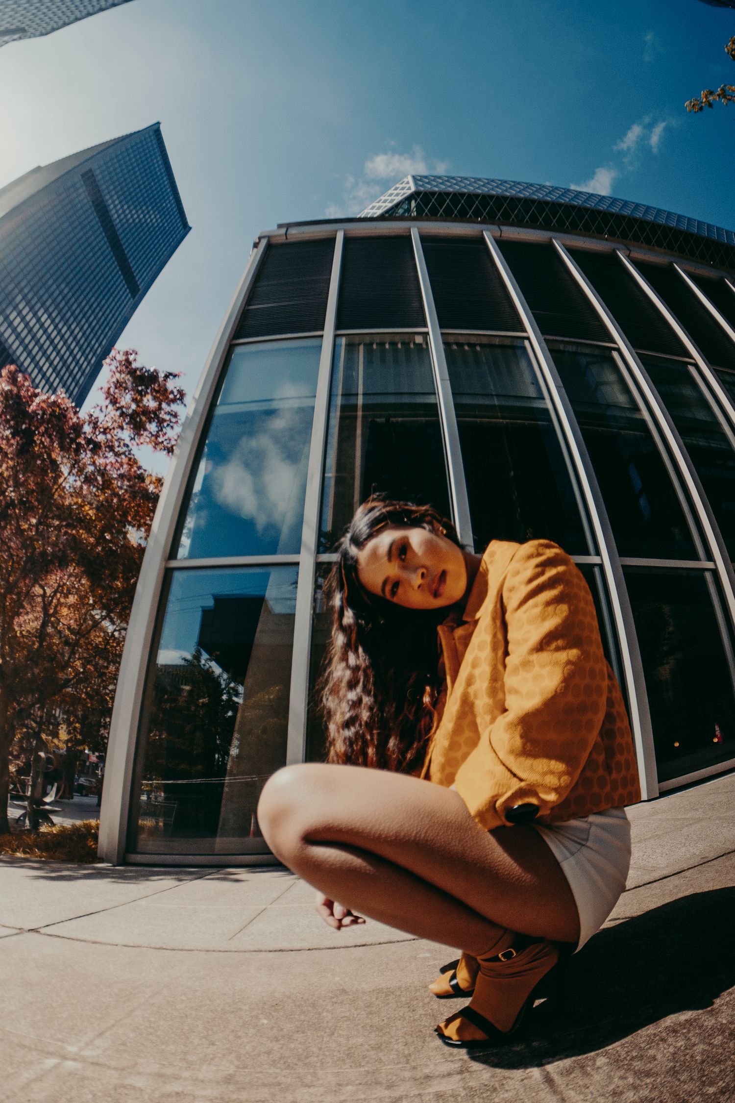 Woman in yellow jacket and white shorts crouches beside a curved glass building with skyscrapers above her.
