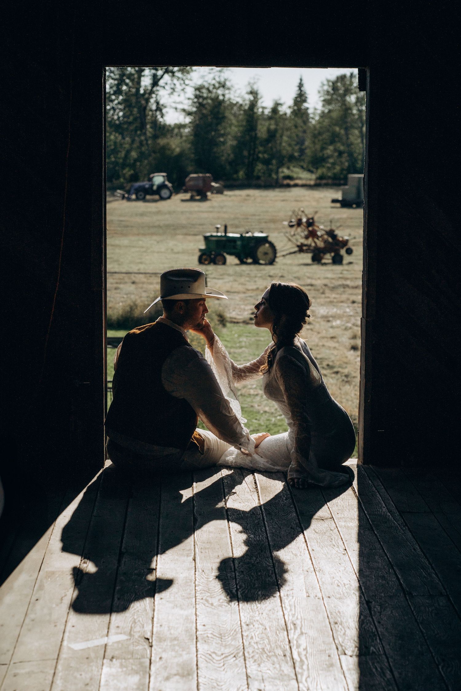 Bride and groom sitting in a barn doorway with farm tractors and fields visible behind them.